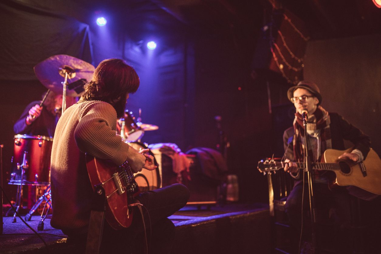 Two male artists seated and playing guitar facing each other in an intimate off Broadway live music venue